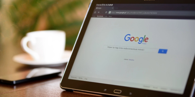 Googled for subconscious mind - Ann Marie MD Tablet on a wooden desk displaying the Google search homepage with a blurred white coffee cup in the background.