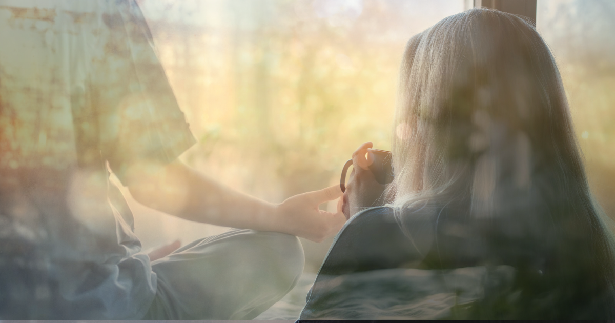 Person with long hair seen from behind sits by a sunlit window as another person hands them a mug.