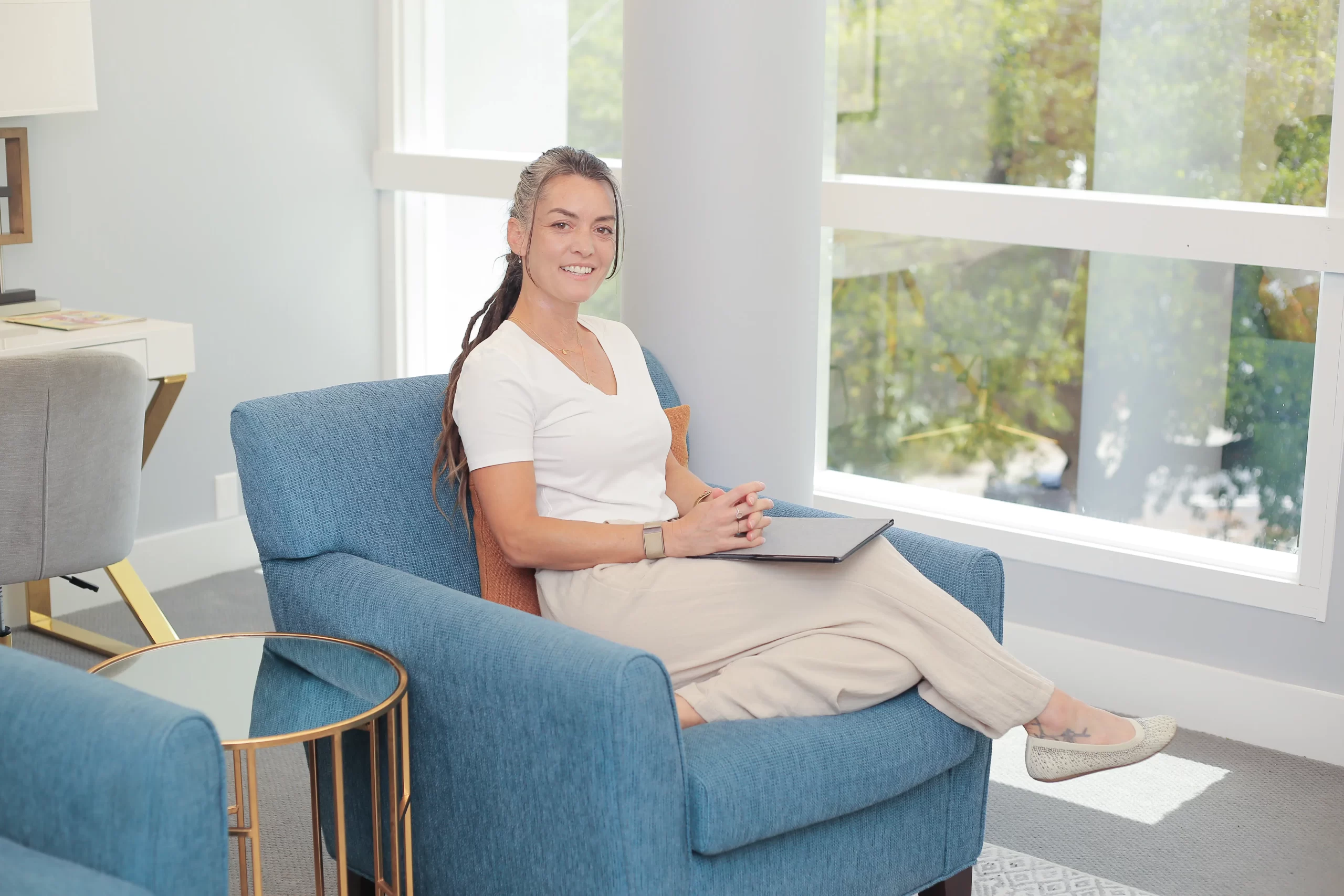 Smiling woman in a white shirt sits on a blue chair in a bright, modern lounge with a notebook on her lap.