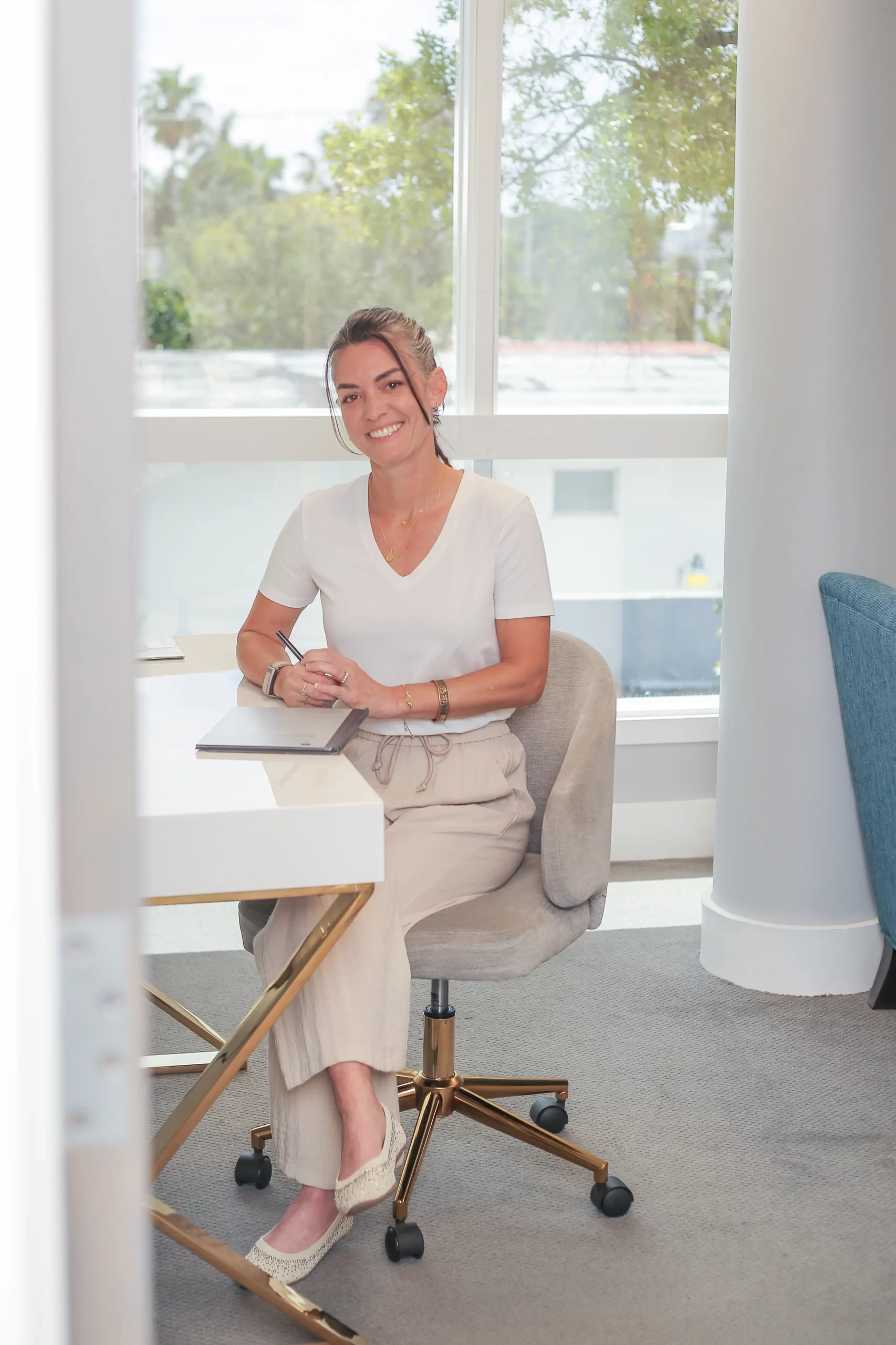 Smiling woman seated at a white desk in a bright office, writing in a notebook.